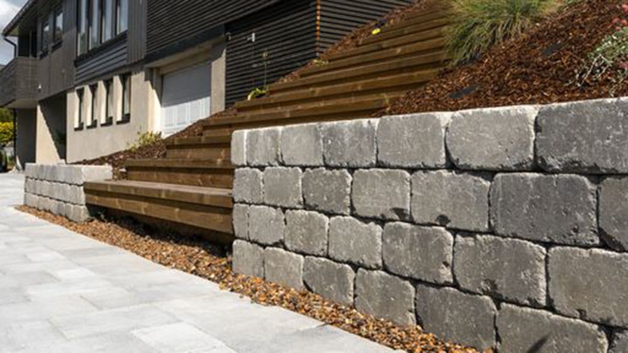 Multiblokk kubbemur som støtter opp skråning foran sort hus.