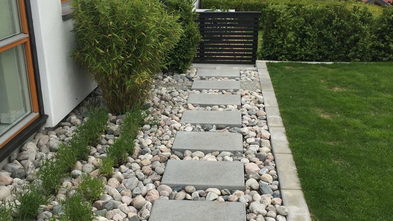 Steinheller og store pyntestein som danner en sti mellom husvegg og plen i en hage.