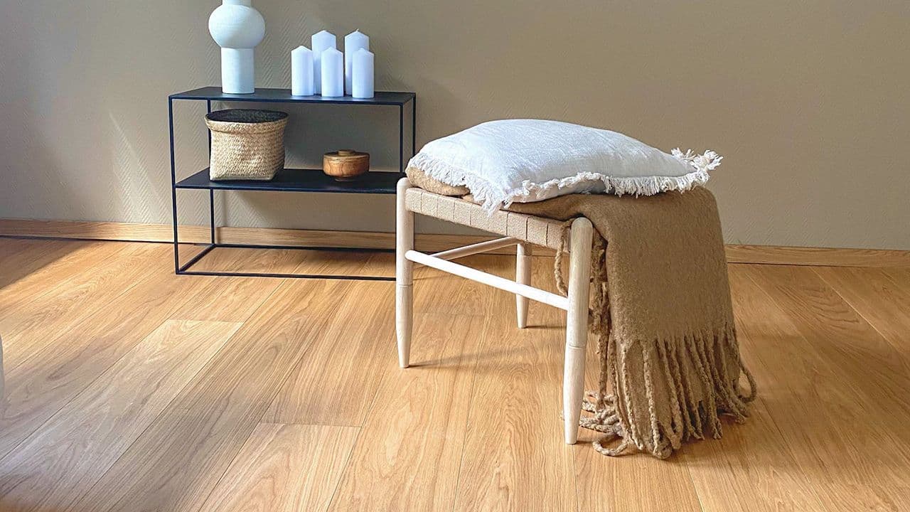 Parkett i gyllen eik med matchende listverk, svart hylle inntil veggen og flettet trebenk med pute og pledd foran.