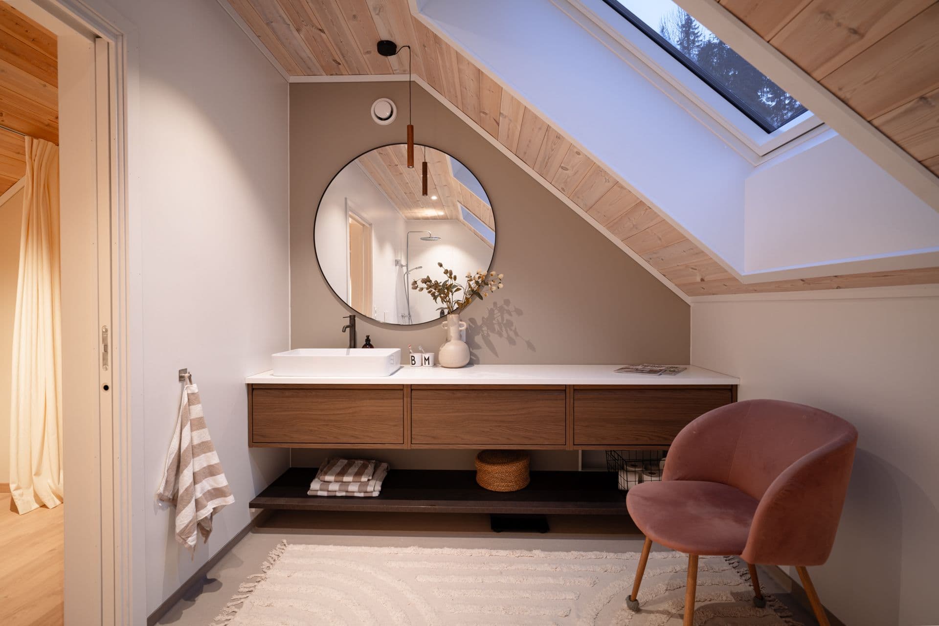 Baderom med baderomsmøbler i mørk eik, rundt speil og stol med rosa stoff.