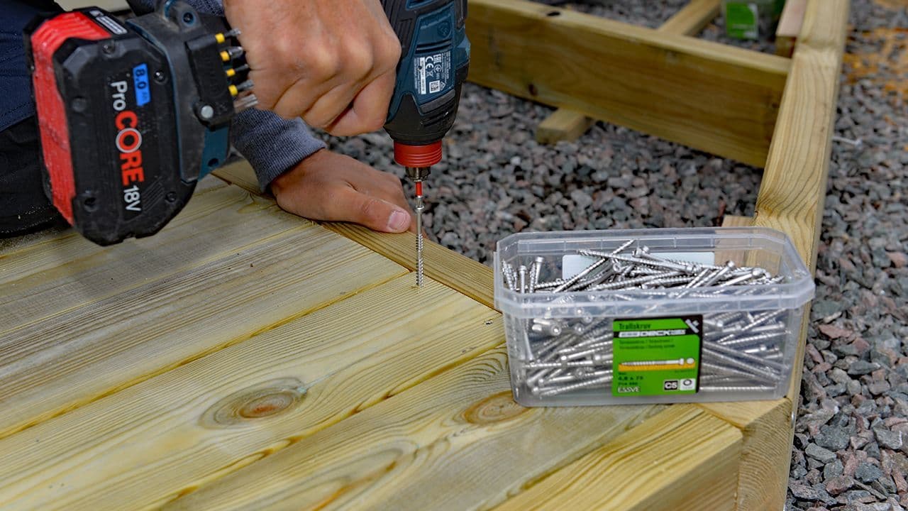 Hender som holder en drill for å bygge terrasse over grus.