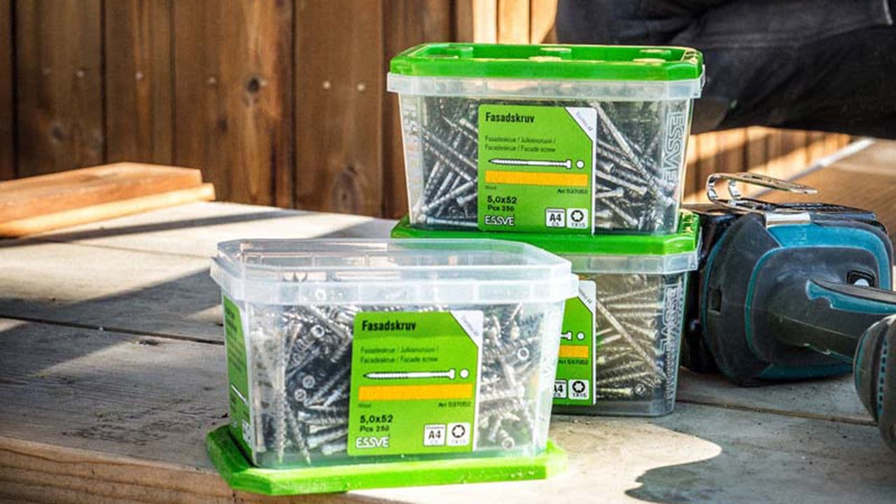 Tre bokser med fasadeskruer som ligger ved siden av en drill på terrassebord.
