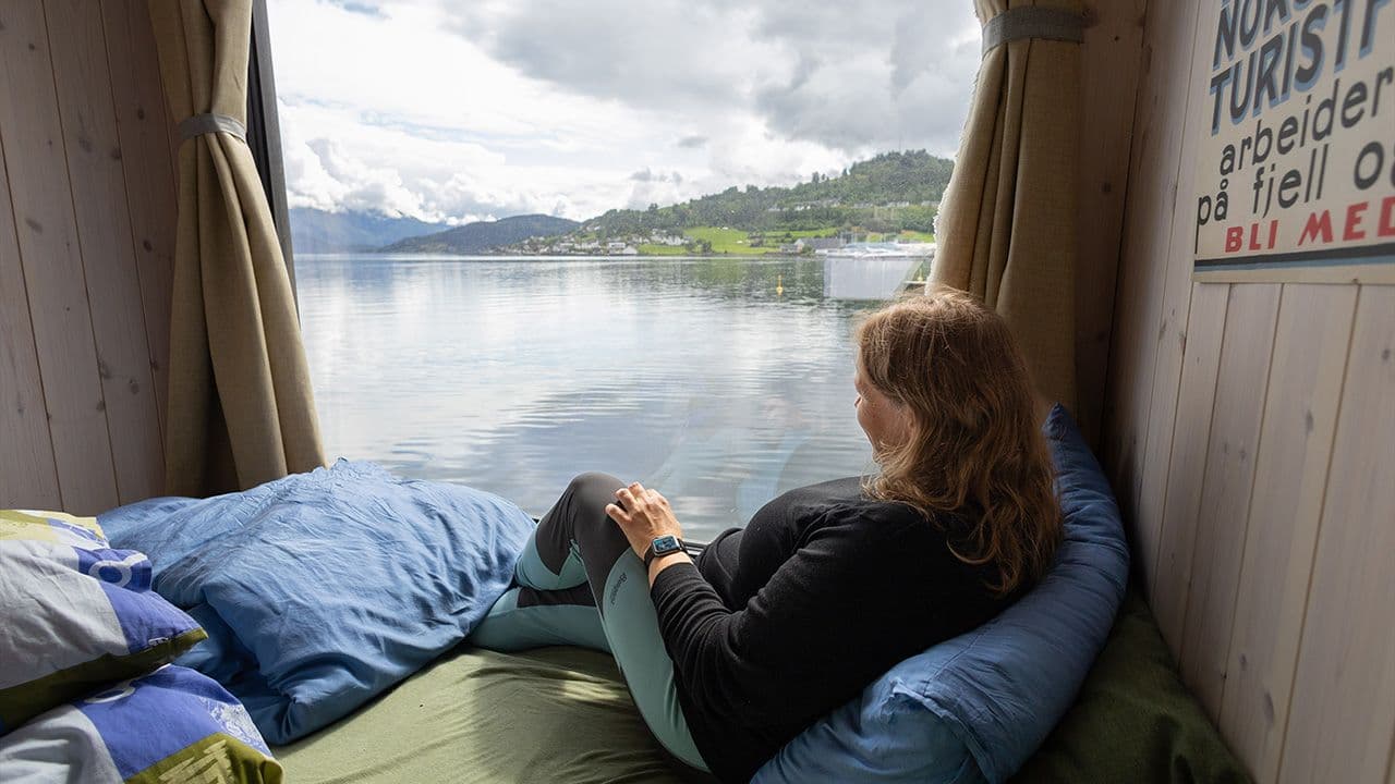 Dame som sitter i en seng i minihytte med utsikt ut over havet.